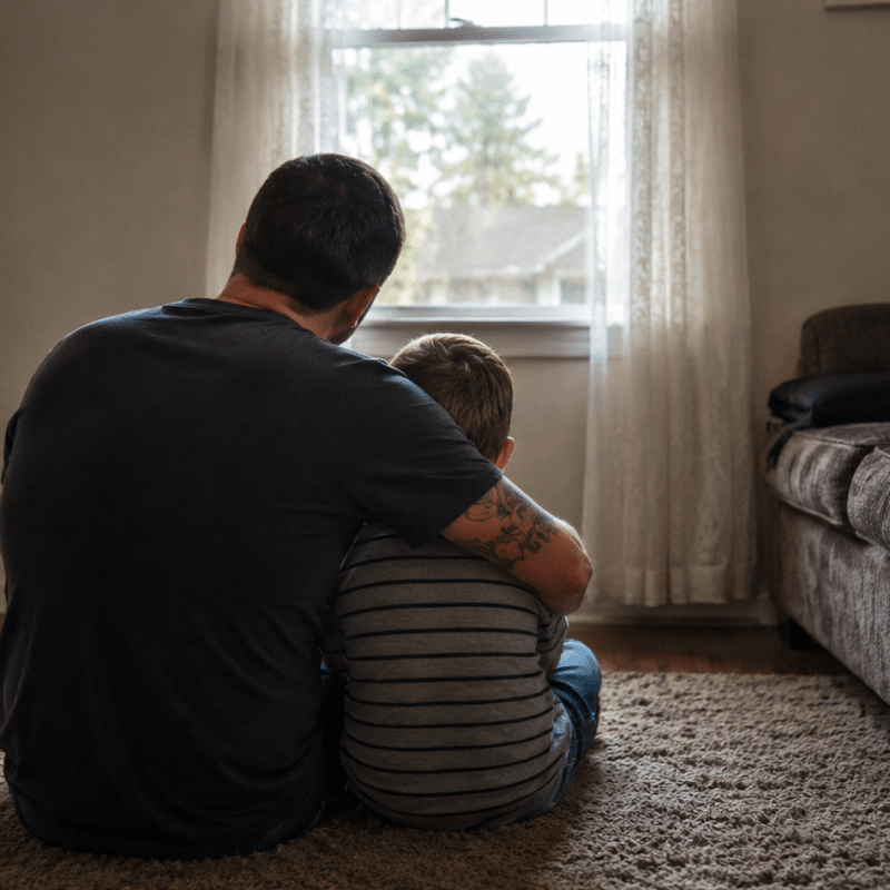 Father and young child sitting together on a living room rug, facing a window in a quiet home setting.